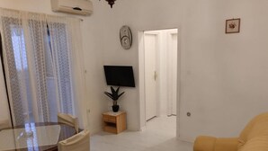 Ett vardagsrum med en gul soffa, ett matbord med stolar, en tv på ett tv-bord och en dörr som leder till ett annat rum.
