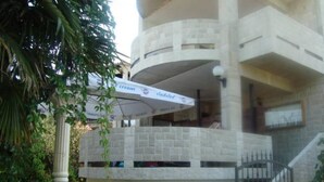 Um edifício de vários andares com fachada de pedra, uma área externa coberta com guarda-sóis e uma escadaria que leva a um nível superior.