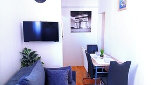 Um espaço de estar compacto com sofá, mesa de jantar com cadeiras, televisão de tela plana, planta em vaso e um quadro na parede.