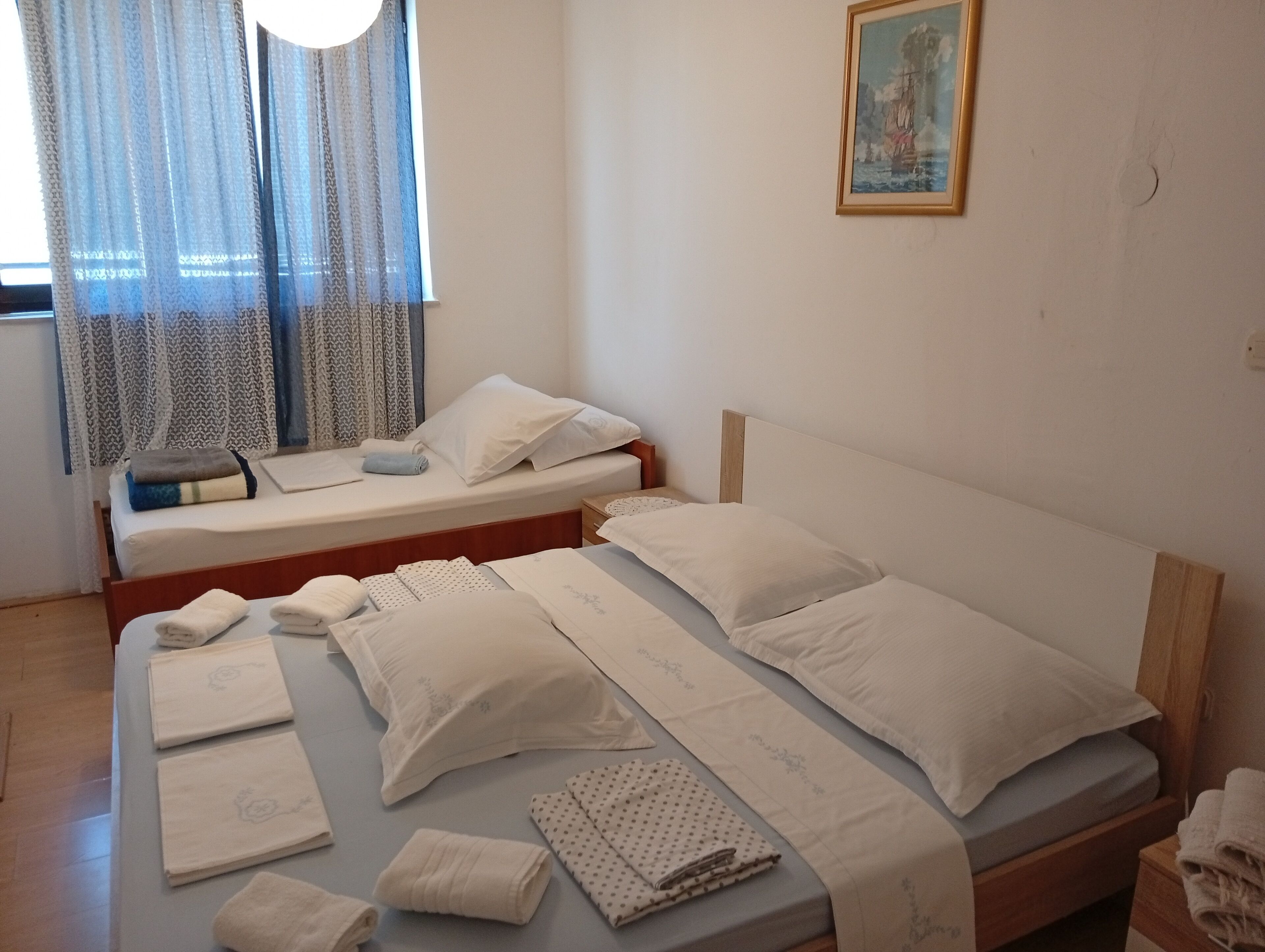 Ein Schlafzimmer mit zwei Betten, einem Fenster mit gemusterten Vorhängen, einem Bild an der Wand und ordentlich drapierten Handtüchern und Kissen auf den Betten.