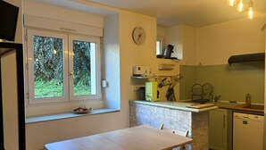 A compact kitchen with a dining area, a window with a view of greenery, a clock on the wall, and basic amenities.