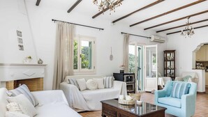 A spacious living room with a white sofa, a blue armchair, and a wooden coffee table. There is a fireplace with a mantel, a chandelier, and a large window with curtains. - Villa Augusta - Two Bedroom Villa, Sleeps 4 (Sporades)