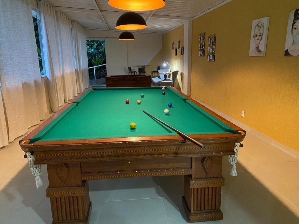 A pool table in a room with a yellow wall, a ceiling light, and a window with curtains.