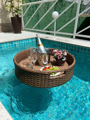 Une scène au bord de la piscine, avec un panier en osier contenant une bouteille de champagne, des verres, des fraises, des raisins et un arrangement floral.