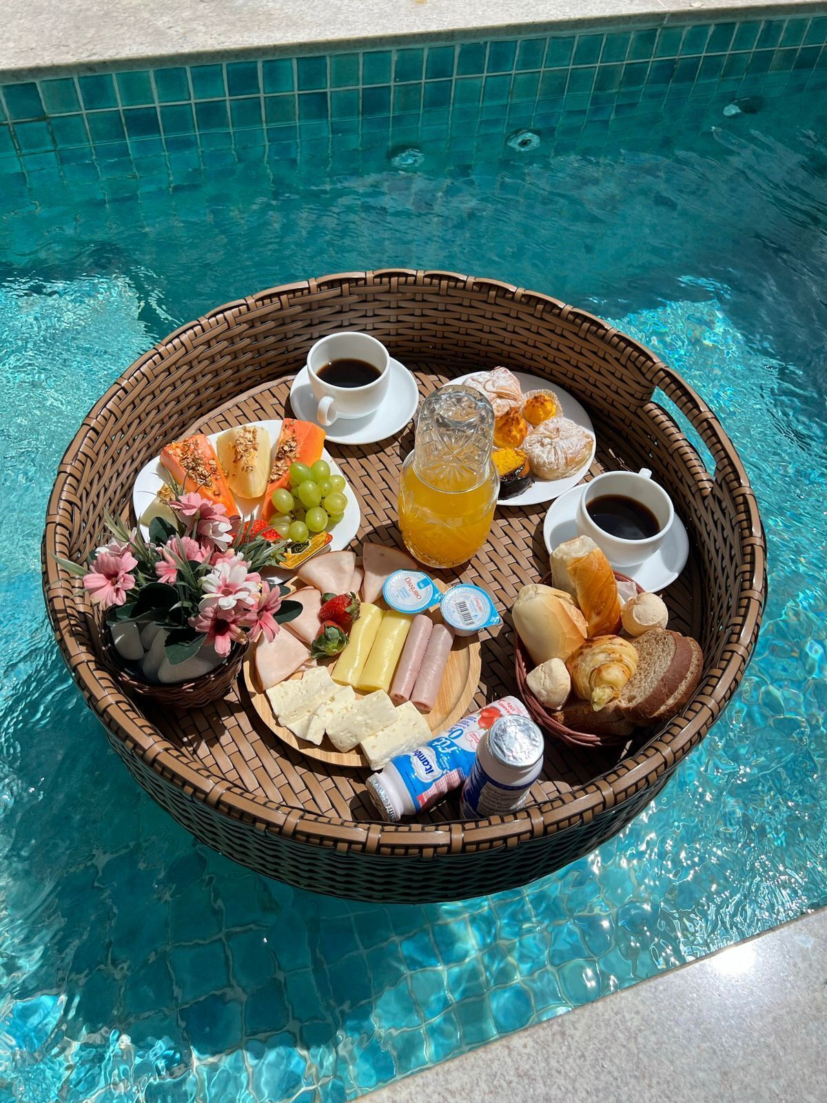 Bandeja de vime com itens de café da manhã, incluindo frutas, doces e bebidas, ao lado de uma piscina.