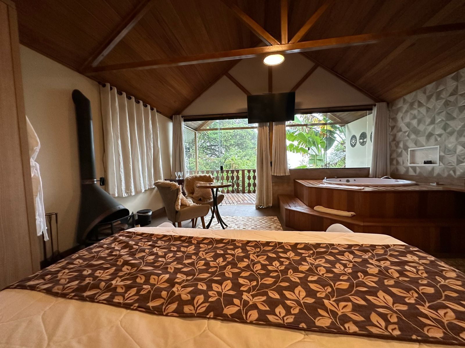 Un dormitorio con una cama grande, techo de madera, ventana con cortinas, una mesita y un baño con bañera y lavabo.