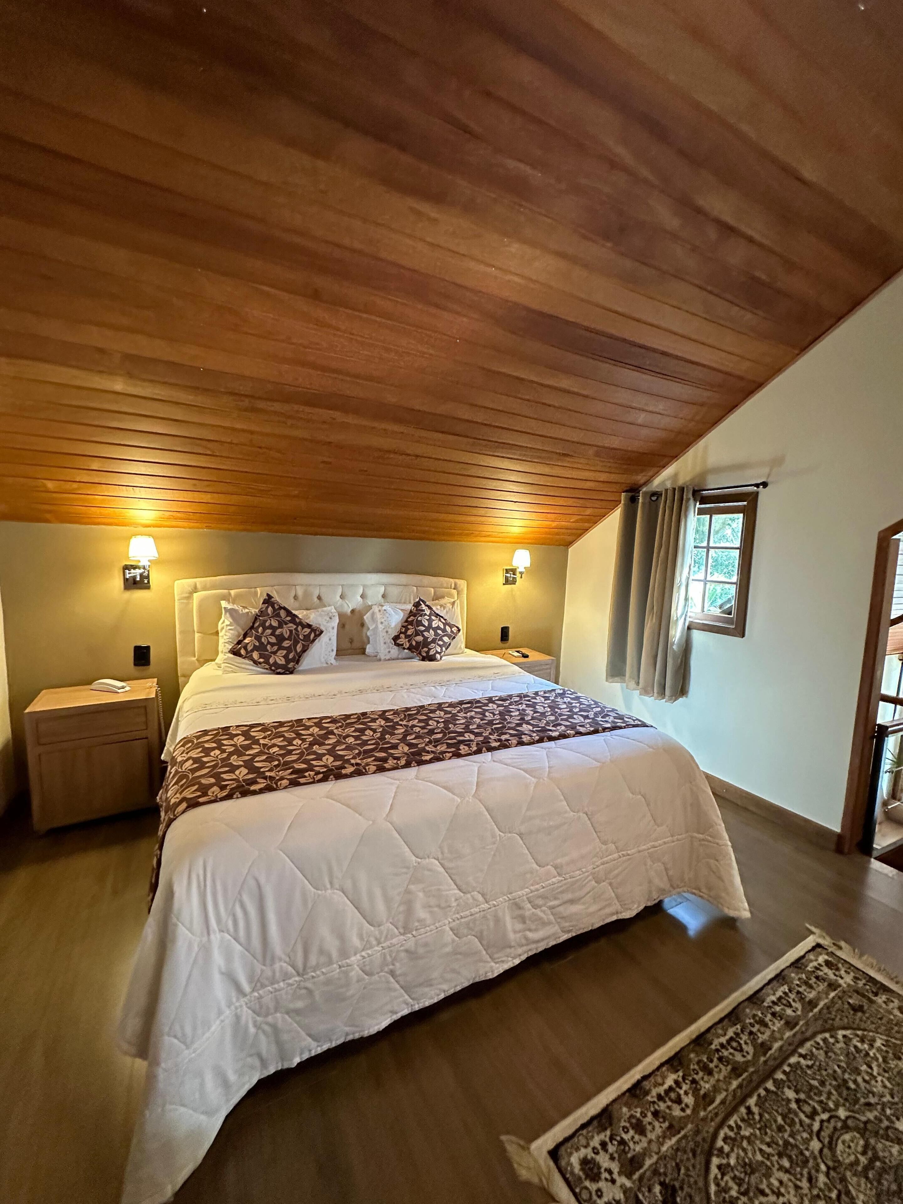 Un dormitorio con techo de madera, una cama grande con edredón blanco acolchado, almohadas estampadas y dos lámparas de noche.