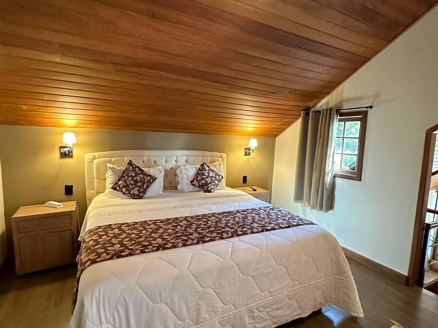 Un dormitorio con techo de madera, una cama grande con edredón blanco acolchado, almohadas estampadas y dos lámparas de noche.