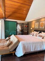 Un dormitorio con cama, sofá, techo de madera, partición de vidrio y pared decorada con dos cuadros de flores.