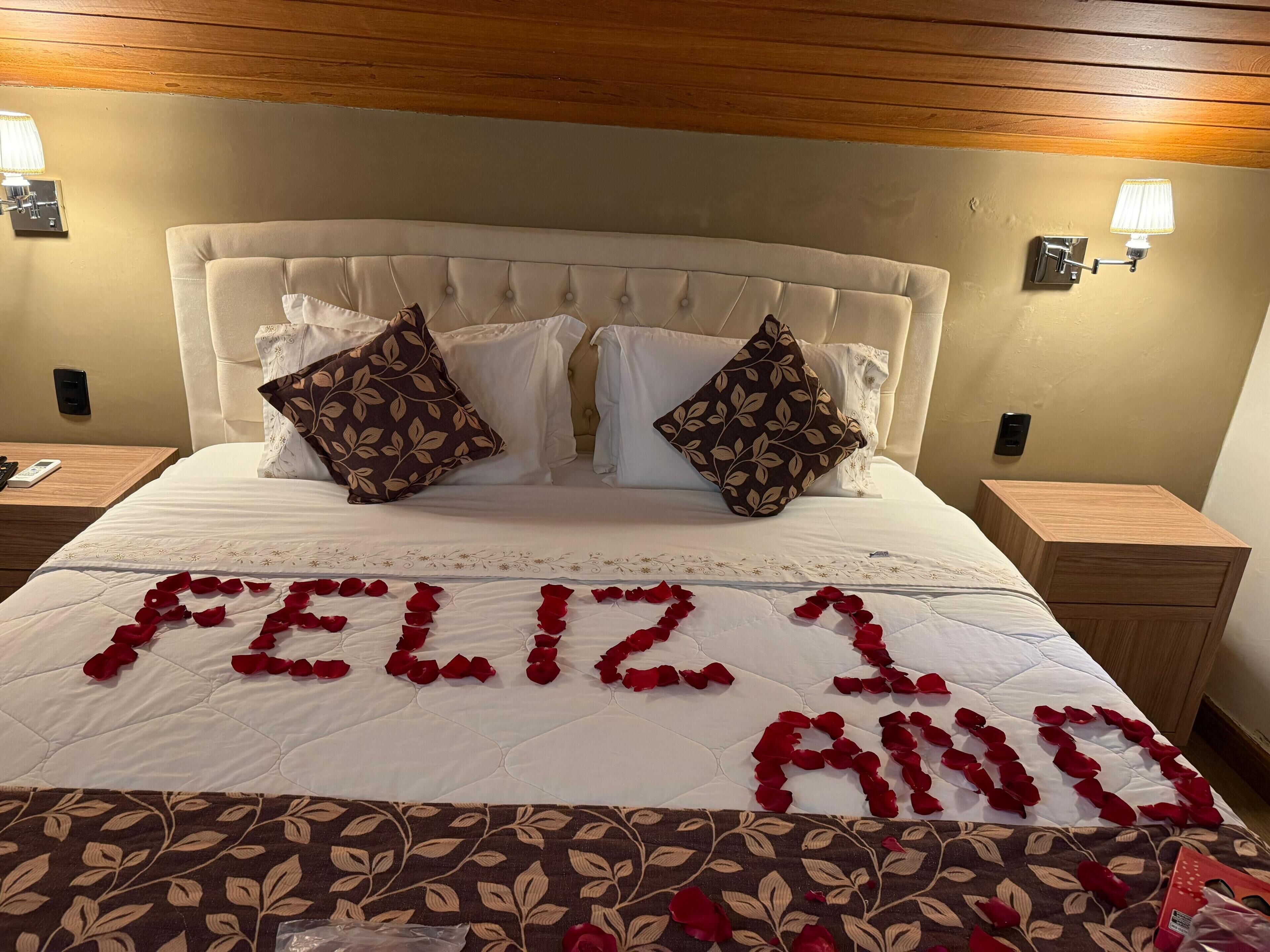 A neatly made bed with a white duvet, two decorative pillows, and a 'Feliz Aniversario' message made of rose petals.