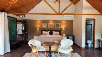 Quarto com uma cama grande, duas poltronas fofas, uma mesinha, um banco e área de chuveiro.