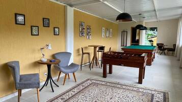 A room with a pool table, a foosball table, a sofa, and a chair. There are framed pictures on the wall and a lamp on a side table.