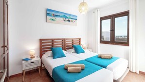 Une chambre d’hôtel avec deux lits, un tableau représentant une scène de plage, une fenêtre avec vue et une lampe de chevet.