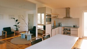 Una cocina moderna con armarios blancos, una campana extractora de acero inoxidable y un horno empotrado. Hay un comedor con mesa y sillas blancas, y una sala de estar con un sofá verde y una planta en maceta.