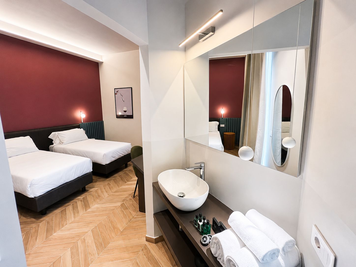 Habitación de hotel con dos camas, un espejo grande, un lavamanos blanco y un suelo con patrón de espiga.