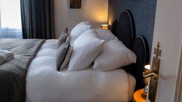 A neatly made bed with white bedding and patterned pillows, a bedside lamp, and a view of the city through a window with curtains.