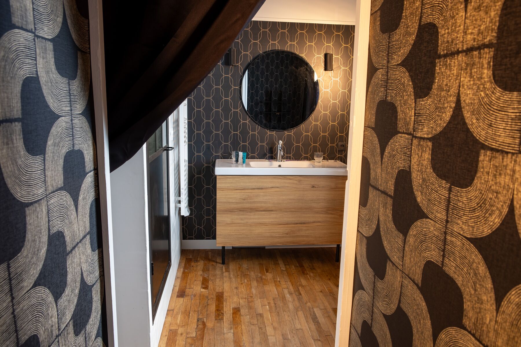 A bathroom with a wooden vanity, a round mirror, and a patterned wall.