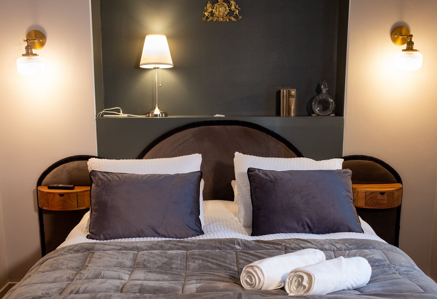 A neatly made bed with a grey quilt, two dark pillows, and a white towel rolled up. There are two bedside tables with lamps, a mirror above the bed, and a decorative item on the wall.