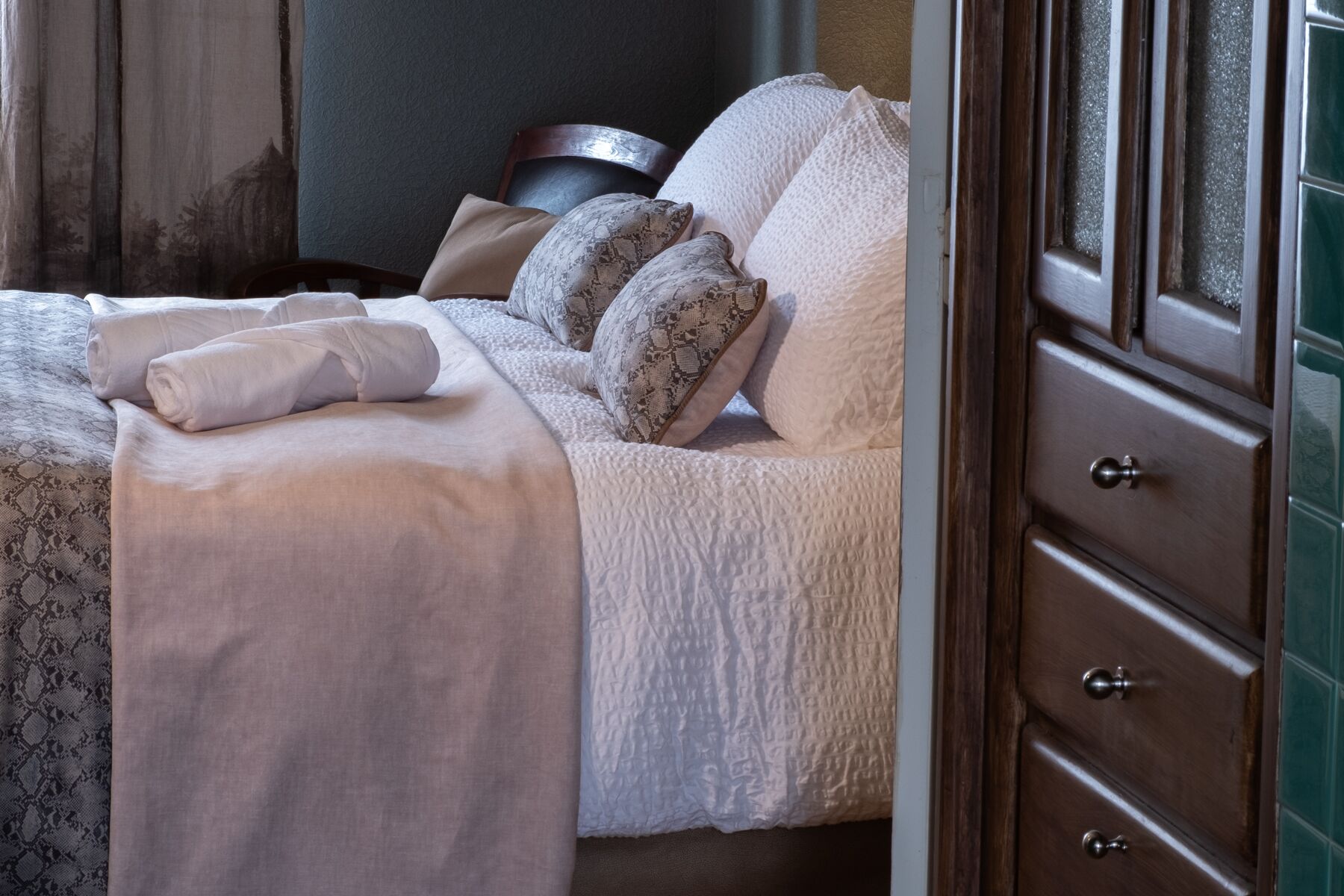 Una cama bien hecha con sábanas blancas y almohadas estampadas, una mesita de noche de madera con lámpara, y un aparador de madera oscura con cajones.
