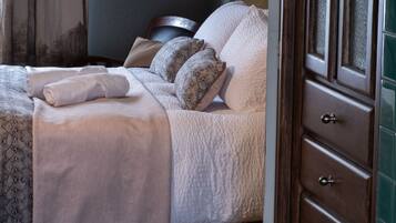 Una cama bien hecha con sábanas blancas y almohadas estampadas, una mesita de noche de madera con lámpara, y un aparador de madera oscura con cajones.