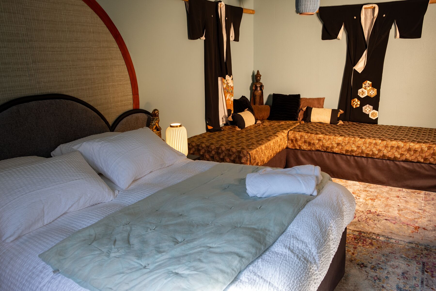 A hotel room with two beds, a large bed in the foreground and a smaller one in the background. There is a wooden headboard on the large bed, and a decorative lamp on a bedside table.