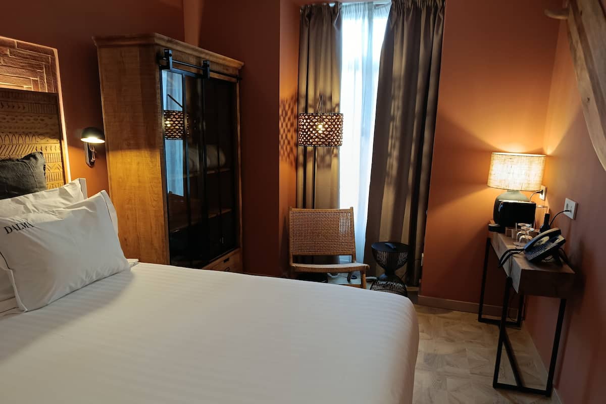 Une chambre d’hôtel comprenant un lit, une armoire, un bureau avec une lampe, une chaise et une fenêtre avec des rideaux.