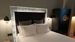 Una cama bien hecha con dos almohadas que dicen "DALMA" en una habitación de hotel con un cabecero estampado, lámparas de noche y un armario de color oscuro.
