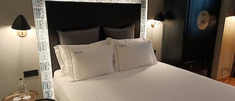 A neatly made bed with two pillows labeled "DALMA" in a hotel room with a patterned headboard, bedside lamps, and a dark-colored wardrobe.