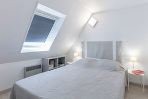 Un dormitorio con techo inclinado, tragaluces, cama con cabecera, mesitas de noche con lámparas y una pequeña estantería con libros.