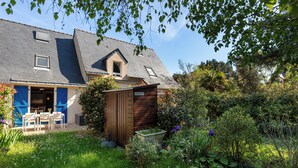 Une maison avec un toit en pente, un petit abri de jardin et un jardin où poussent différentes plantes et fleurs.