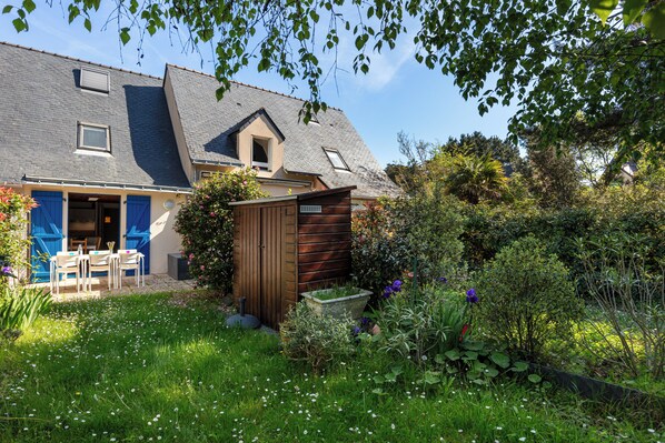 Una casa con techo inclinado, un cobertizo pequeño y un jardín con diversas plantas y flores.