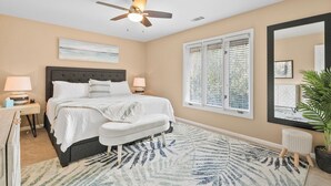 A bedroom with a bed, bedside lamps, a ceiling fan, a window with blinds, a large mirror, and a potted plant.