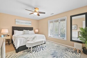 Une chambre à coucher avec un lit, des lampes de chevet, un ventilateur de plafond, une fenêtre avec des stores, un grand miroir et une plante en pot.