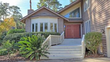 A two-story house with a front porch, a red door, and a small front yard with a palm tree and shrubs.