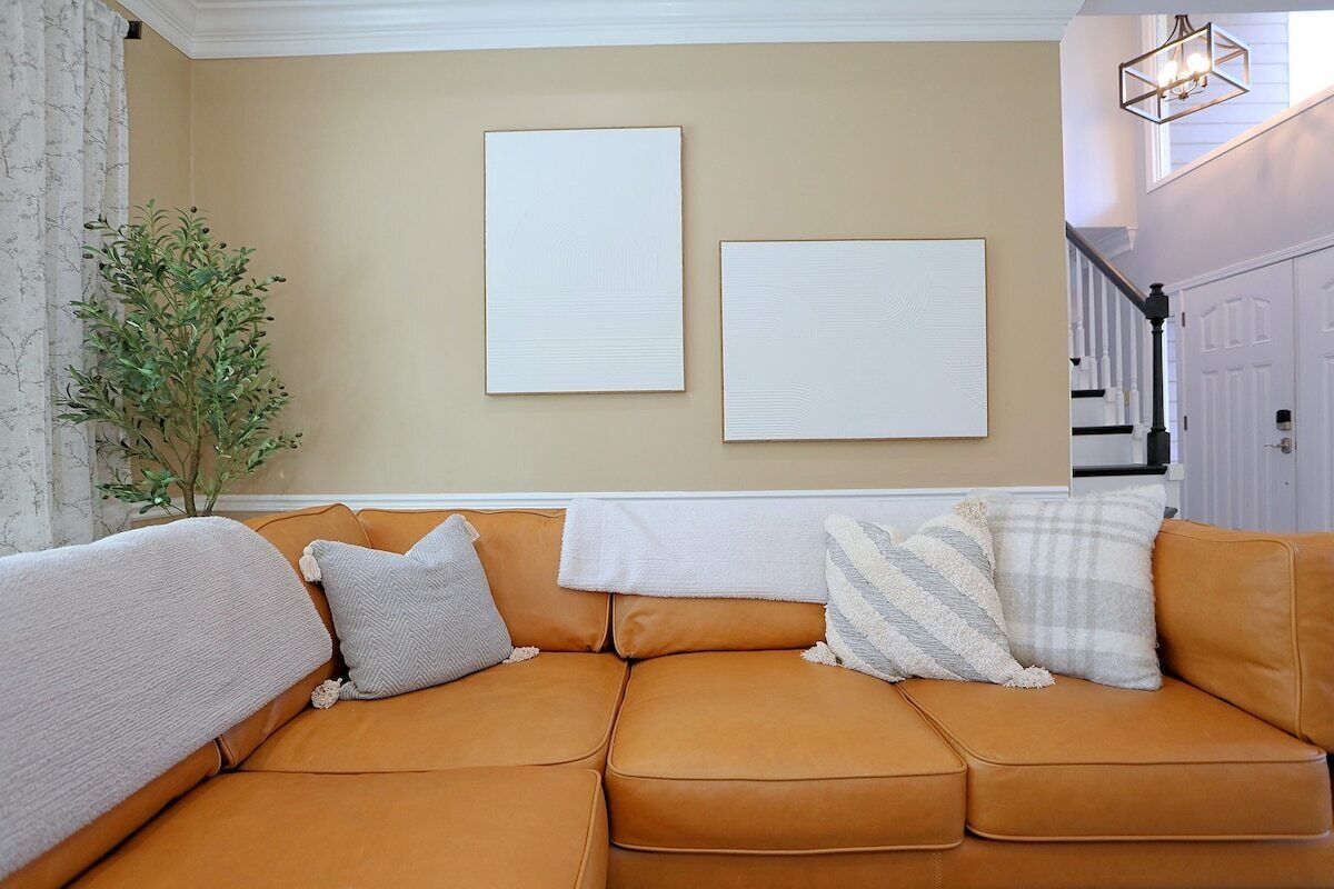 A living room with a tan leather sofa, two framed artworks on the wall, a potted plant, and a staircase in the background.