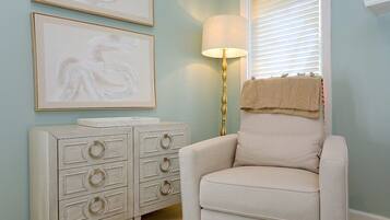 Une pièce comprenant un fauteuil blanc, une commode blanche avec des tiroirs, une lampe et deux œuvres d’art encadrées accrochées au mur.