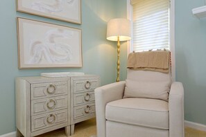 Een kamer met een witte fauteuil, een wit dressoir met lades, een lamp en twee kunstwerken in een lijst aan de muur.