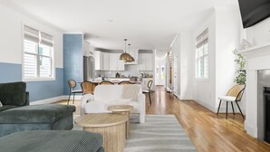 A spacious living area with a sofa, coffee table, and dining table. A kitchen is visible in the background. - Boho Luxe Stay Game Room, Fire Pit & Playground (Raleigh)