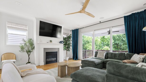A living room with a fireplace, a television, a ceiling fan, and a large window with curtains. - Boho Luxe Stay Game Room, Fire Pit & Playground (Raleigh)