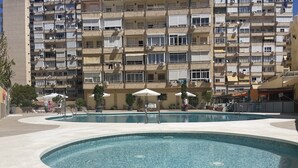 Ein Schwimmbecken mit umlaufender Promenade, ein mehrstöckiges Gebäude mit Balkonen und Außenbereichen zum Sitzen.