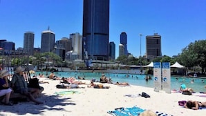 A beach with people sunbathing and swimming, a city skyline in the background, and a sign with symbols. - Lovely 2 Beds Apt with City View at South Brisbane (South Brisbane)