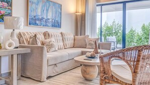 A modern living room with a beige sofa, a round coffee table, and a wicker chair. A painting of sailboats hangs on the wall, and a large window offers a view of greenery outside. - ENJOY THIS BEAUTIFUL APARTMENT IN PUNTA CANA (Higüey)