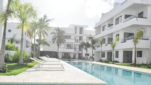 A swimming pool with lounge chairs, palm trees, and modern buildings in the background. - ENJOY THIS BEAUTIFUL APARTMENT IN PUNTA CANA (Higüey)