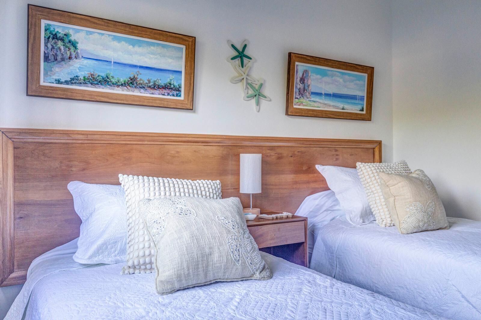 Habitación de hotel con dos camas, ropa de cama blanca, cabecera de madera, mesita de noche con lámpara y dos cuadros enmarcados en la pared.