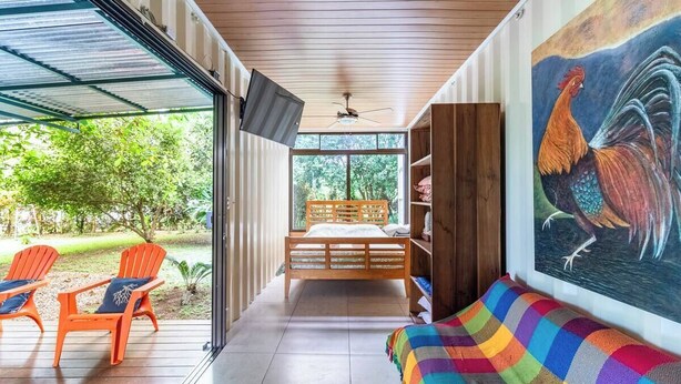 A room with a bed, a colorful couch, a ceiling fan, a large window, and a painting of a rooster.