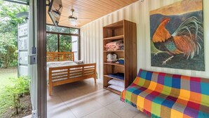 A room with a bed, a wooden shelf with folded clothes, a colorful blanket, and a painting of a rooster.