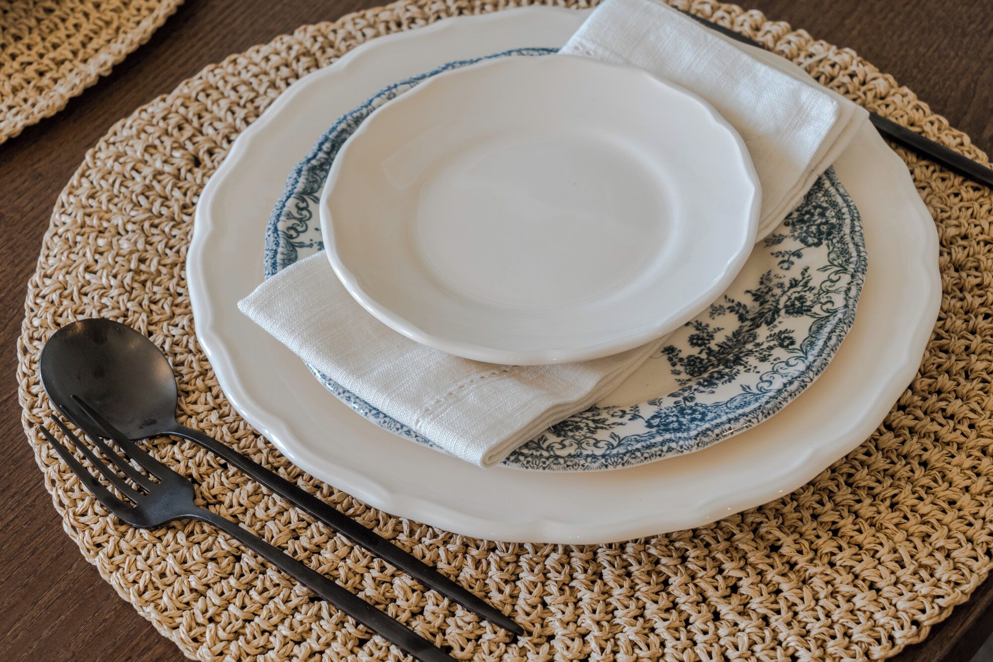 Une table dressée avec un set de table en fibres tressées, une assiette blanche ornée d’un motif bleu, une serviette blanche pliée, une fourchette noire et une cuillère.