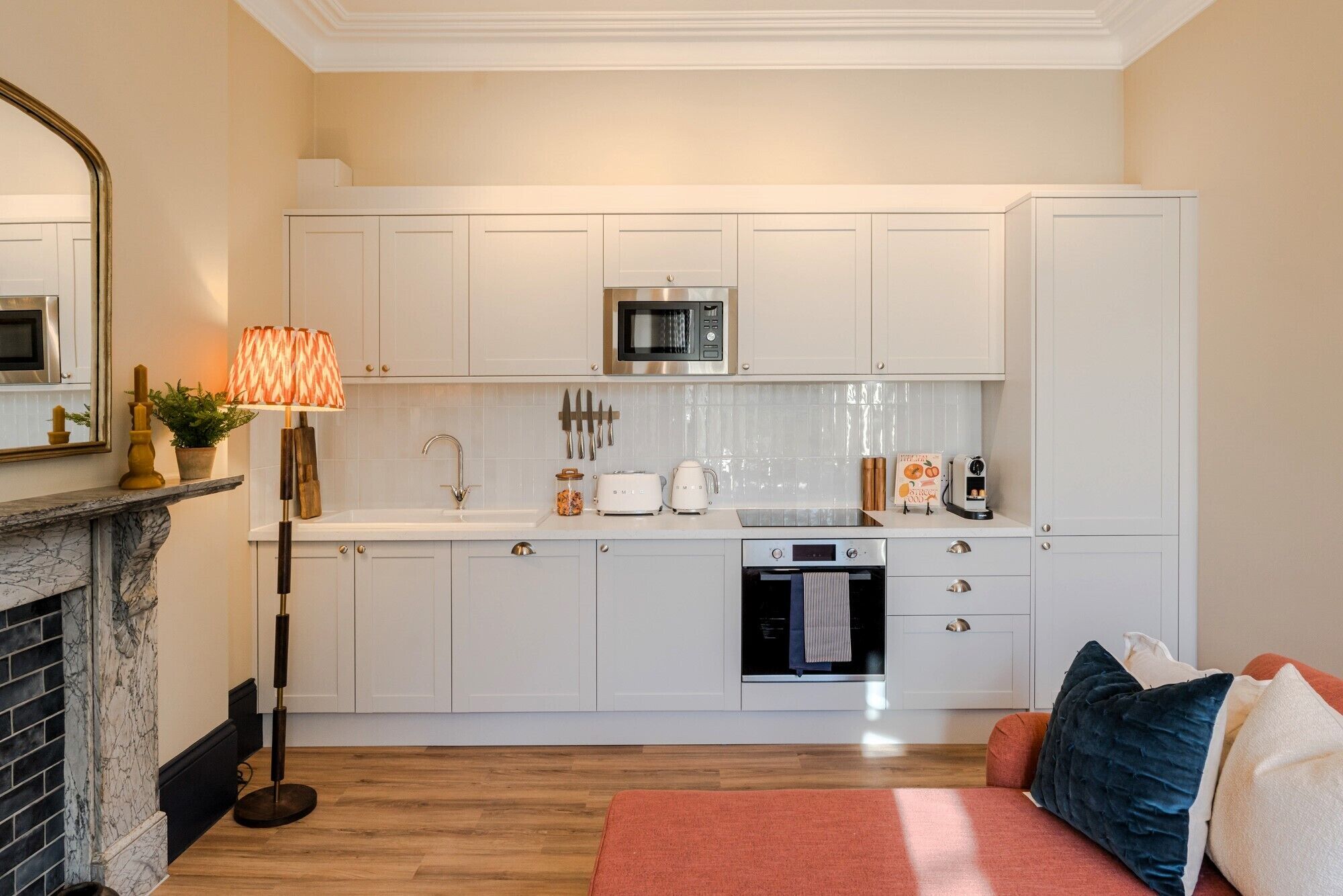 A modern kitchen with white cabinets, a microwave, a sink, and a stove. There is a red rug and a couch with pillows in the foreground.