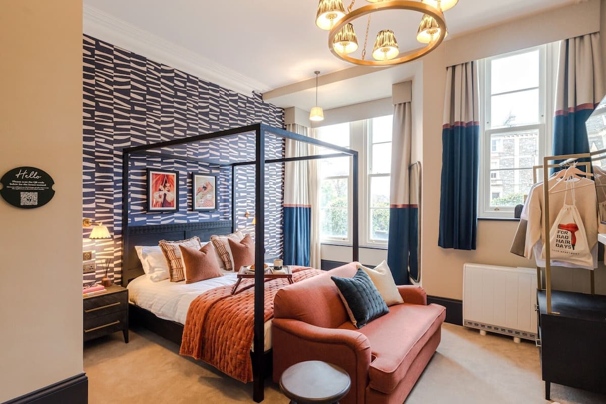 Quarto moderno com uma cama de dossel, um sofá e uma pequena mesa. Há uma janela com cortinas, um radiador e um aviso na parede.