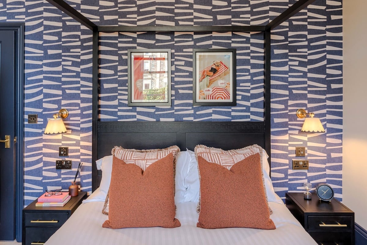 Quarto com parede estampada, duas imagens emolduradas, cama com duas almofadas, mesas de cabeceira com luminárias e um criado-mudo com relógio.
